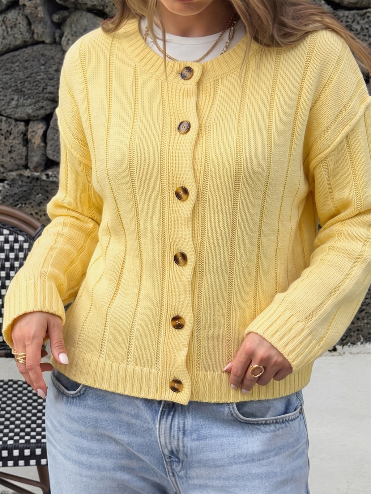 A woman wearing a pale yellow ribbed knit cardigan sweater with brown buttons, paired with light blue jeans.