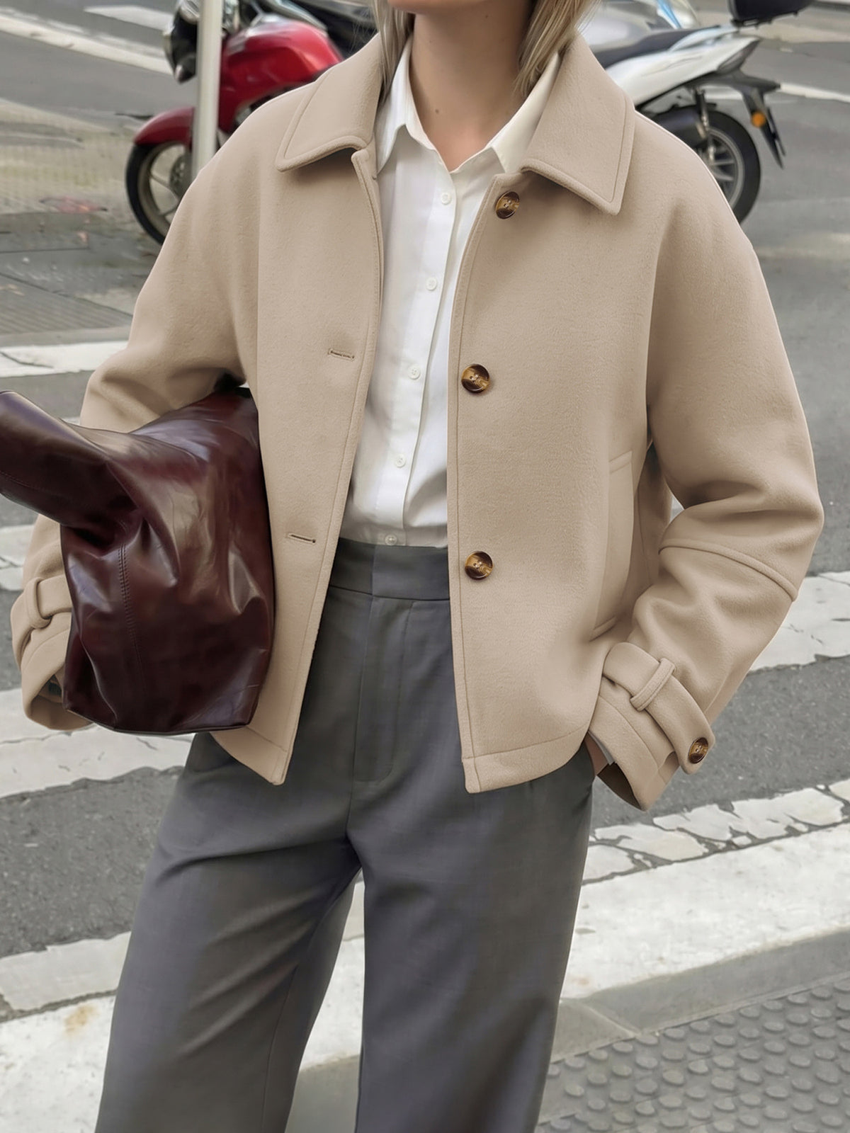 A person wearing a beige cropped jacket over a white button-up shirt and gray trousers, holding a dark brown leather bag on a city street with motorcycles in the background.