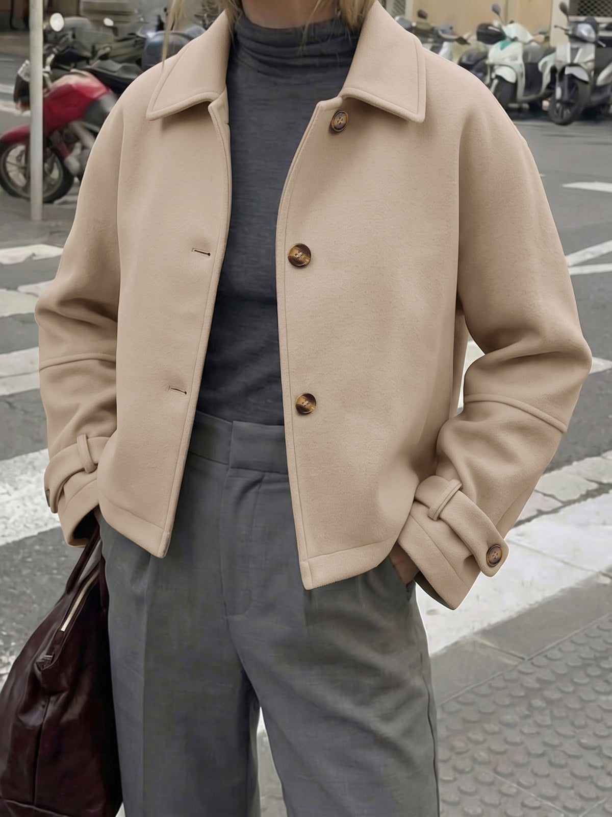 A person wearing a beige cropped jacket over a dark gray turtleneck and gray trousers, standing on a city street with parked motorbikes in the background.