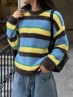 A person wearing a fuzzy crewneck sweater with horizontal stripes in light blue, yellow, and dark brown, paired with light blue jeans and a black shoulder bag.