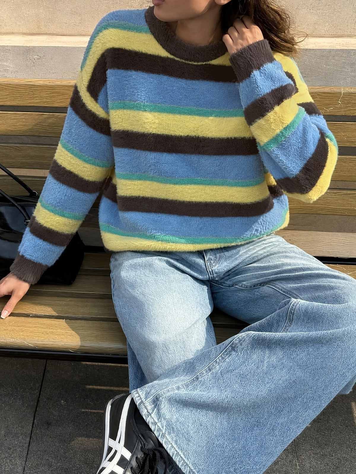 A person sitting on a wooden bench wearing a fuzzy long-sleeve sweater with horizontal stripes in light blue, yellow, dark brown, and teal, paired with light-wash wide-leg jeans and black sneakers.