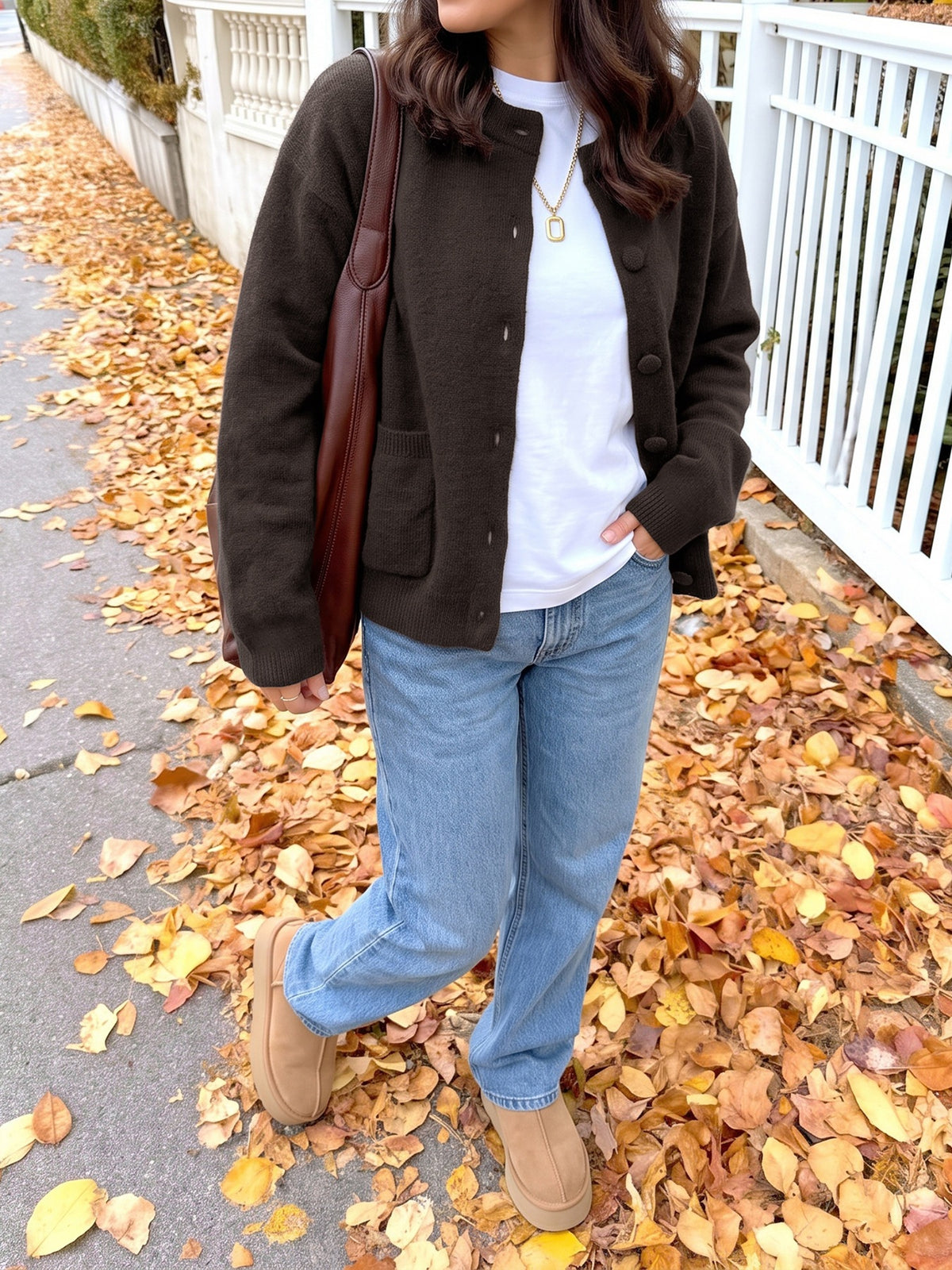 A woman wearing a dark brown button-up cardigan over a white t-shirt, light blue jeans, and tan suede boots, carrying a brown leather shoulder bag while walking on a path covered with fallen autumn leaves.