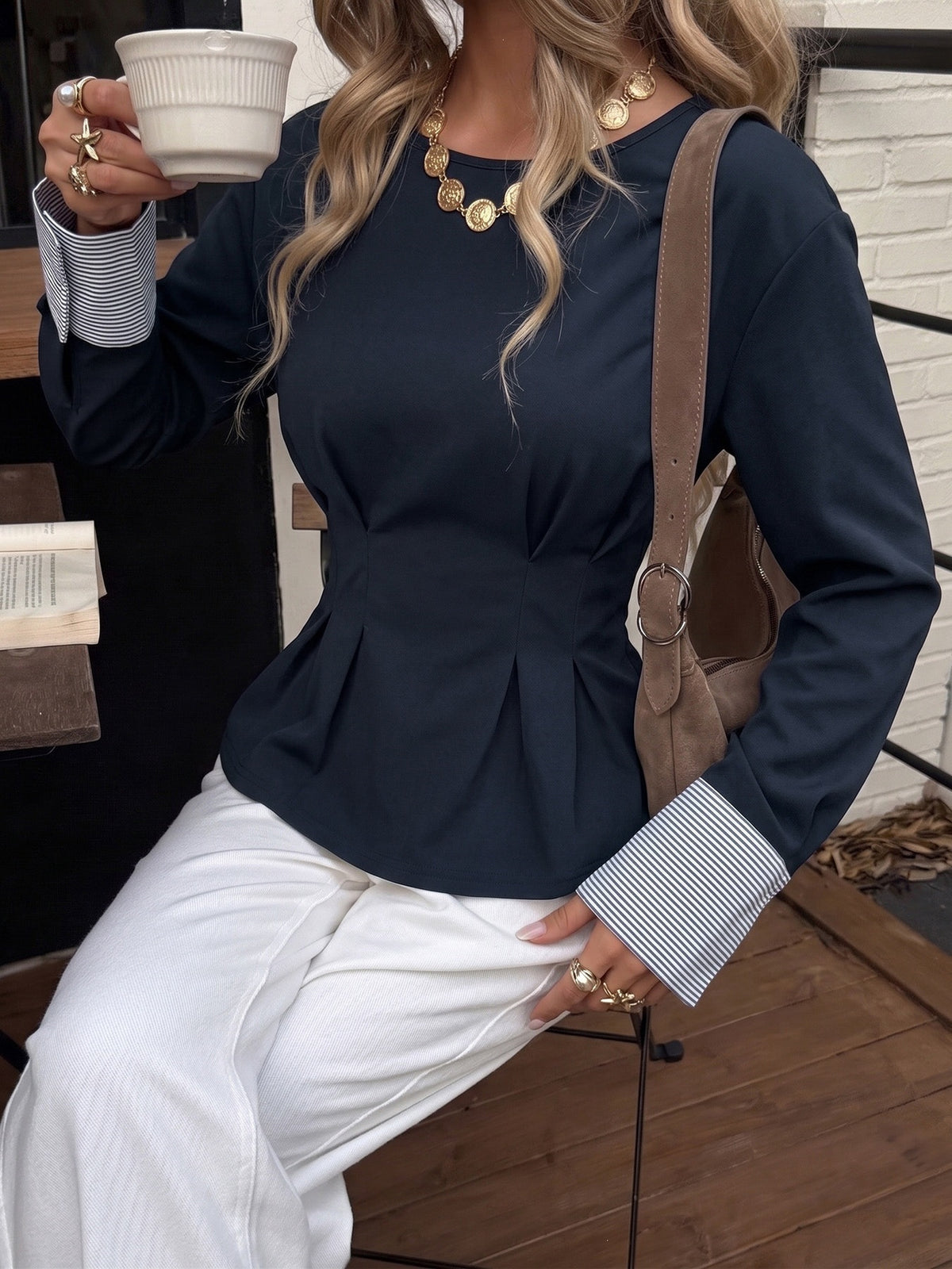 A woman wearing a navy blue long-sleeve peplum top with striped buttoned cuffs, paired with white pants and a brown shoulder bag, holding a white coffee cup.