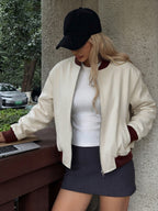A woman wearing a cream-colored bomber jacket with dark red ribbed trim, a white top, a dark gray mini skirt, and a black baseball cap, leaning against a concrete pillar next to a wooden table.