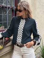 A woman wearing a dark teal long-sleeve blouse with prominent white crochet lace trim down the front and along the sleeves, paired with white pants and sunglasses, standing outdoors next to a window.