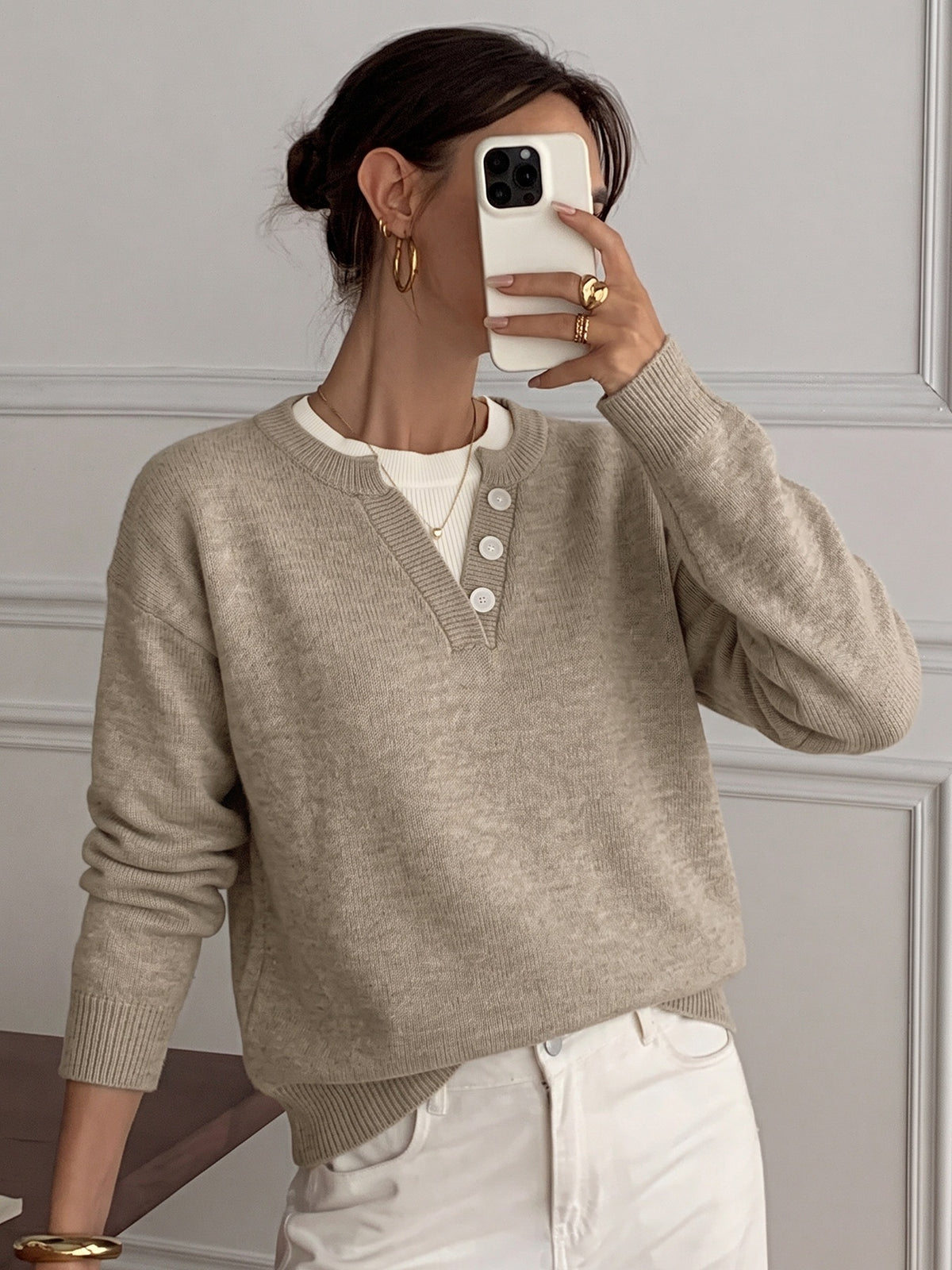 A woman takes a mirror selfie wearing a light beige knit sweater with a V-neck and three white buttons, layered over a white top and paired with white pants.