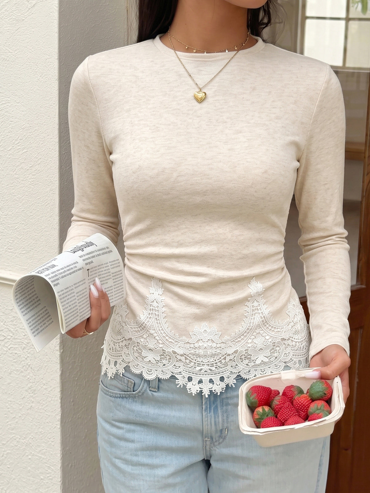 A woman wearing a fitted long-sleeve cream top with white lace trim at the hem, paired with light blue jeans. She is holding a folded newspaper in one hand and a small container of strawberries in the other, standing indoors near a wall and window.