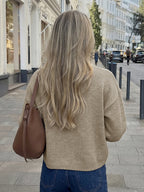 A woman viewed from behind on a city sidewalk, wearing a beige knit sweater and blue jeans, with a brown shoulder bag slung over her shoulder.