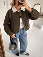 A woman takes a mirror selfie wearing a brown corduroy jacket with a white sherpa collar, paired with wide-leg blue jeans, a white crop top, and black polka-dot flat shoes, holding a black and white polka-dot handbag.