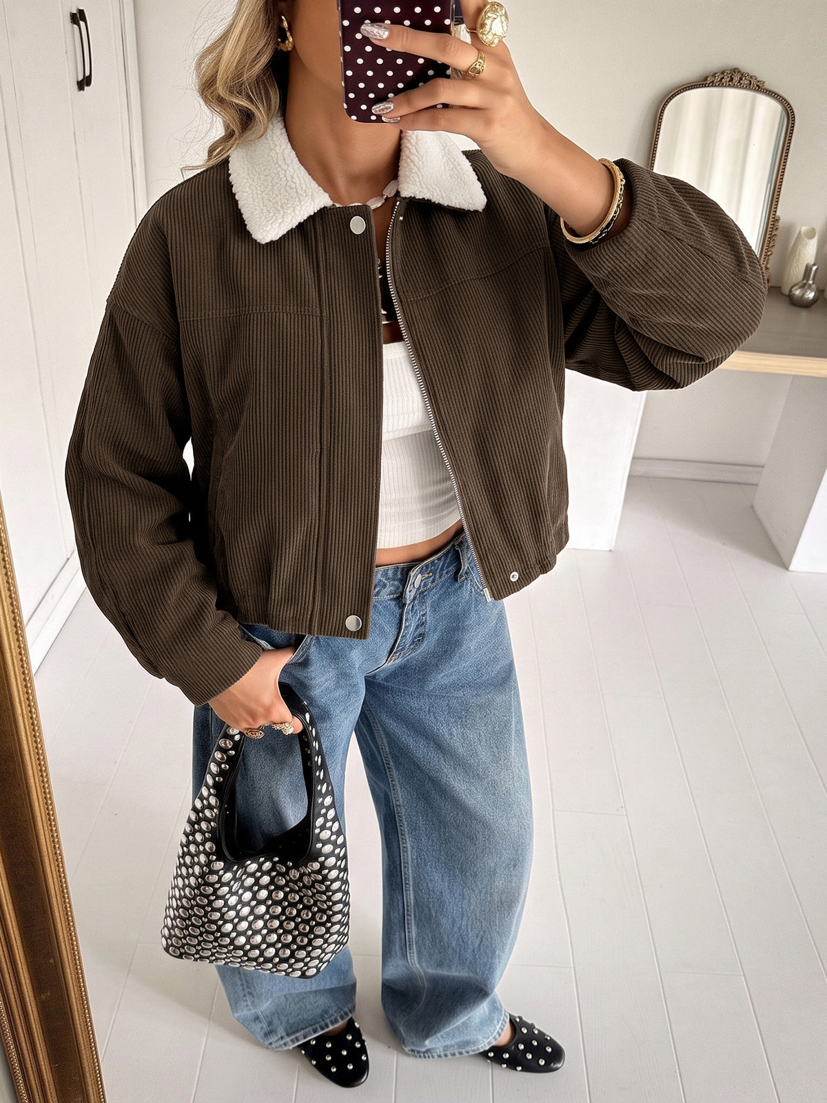 A woman takes a mirror selfie wearing a brown corduroy jacket with a white sherpa collar, paired with wide-leg blue jeans, a white crop top, and black polka-dot flat shoes, holding a black and white polka-dot handbag.