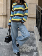 A person wearing a fuzzy knit sweater with horizontal stripes in blue, yellow, and brown, paired with wide-leg light-wash jeans, black sneakers, and carrying a small black handbag while standing outdoors on a city sidewalk.
