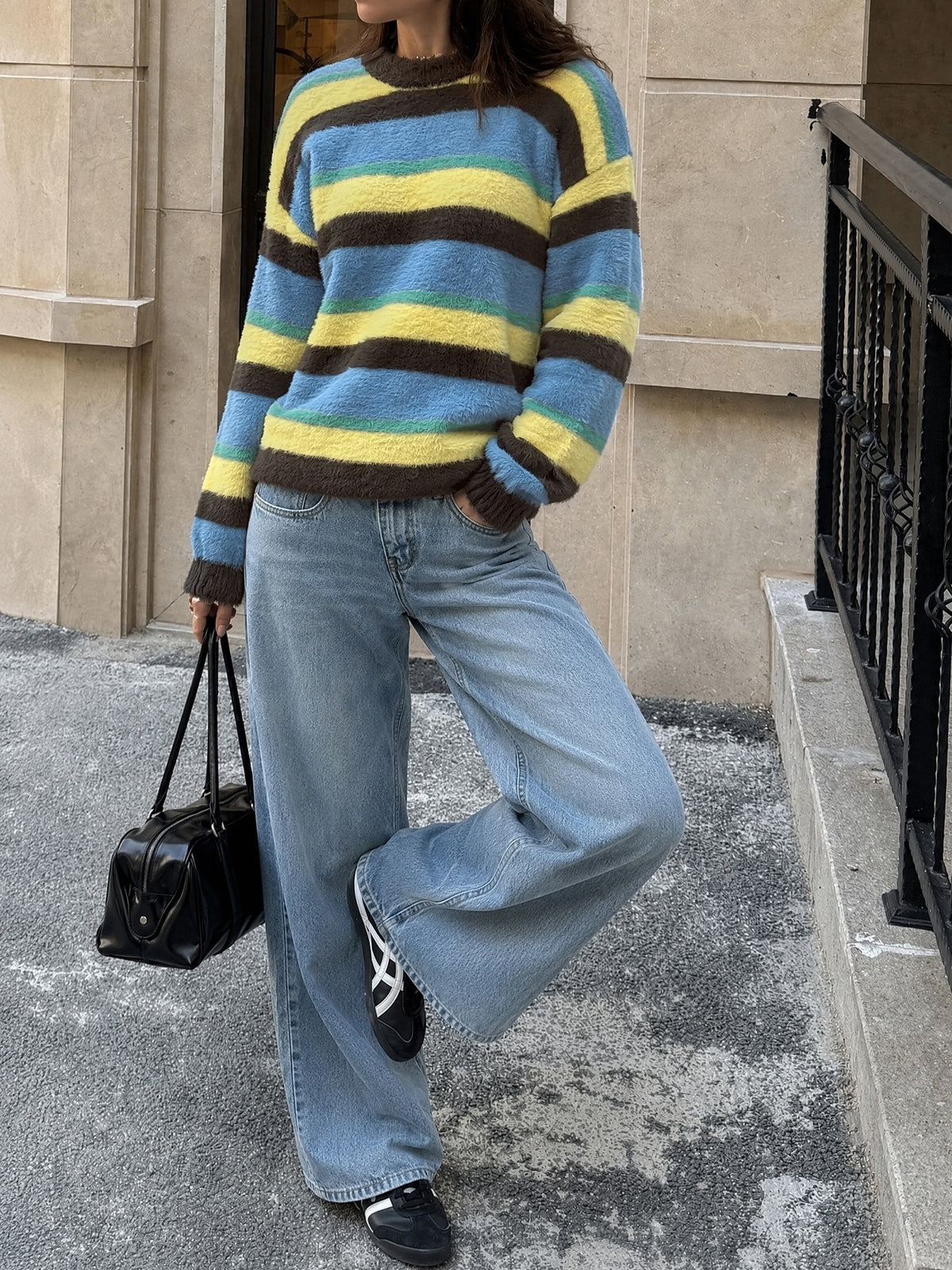 A person wearing a fuzzy knit sweater with horizontal stripes in blue, yellow, and brown, paired with wide-leg light-wash jeans, black sneakers, and carrying a small black handbag while standing outdoors on a city sidewalk.