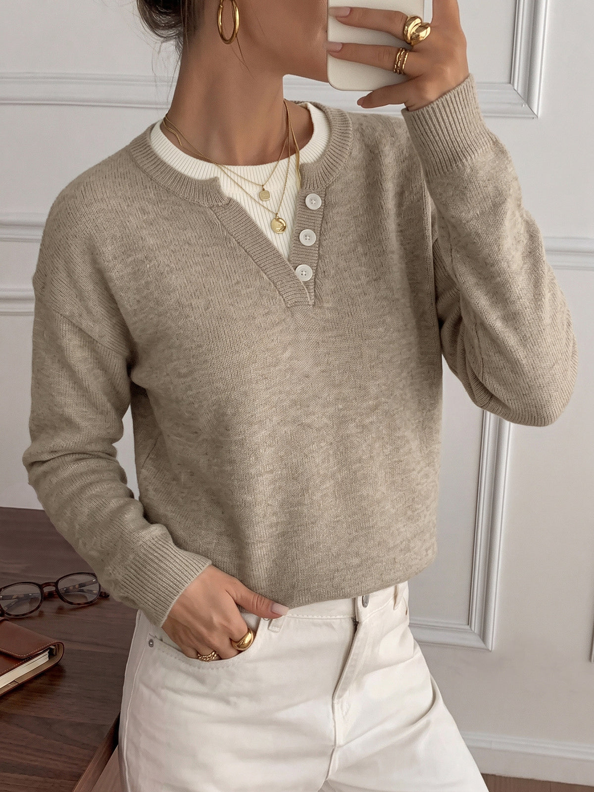 A woman wearing a light taupe knit henley sweater over a cream ribbed shirt and white pants, holding a white phone in front of her face.