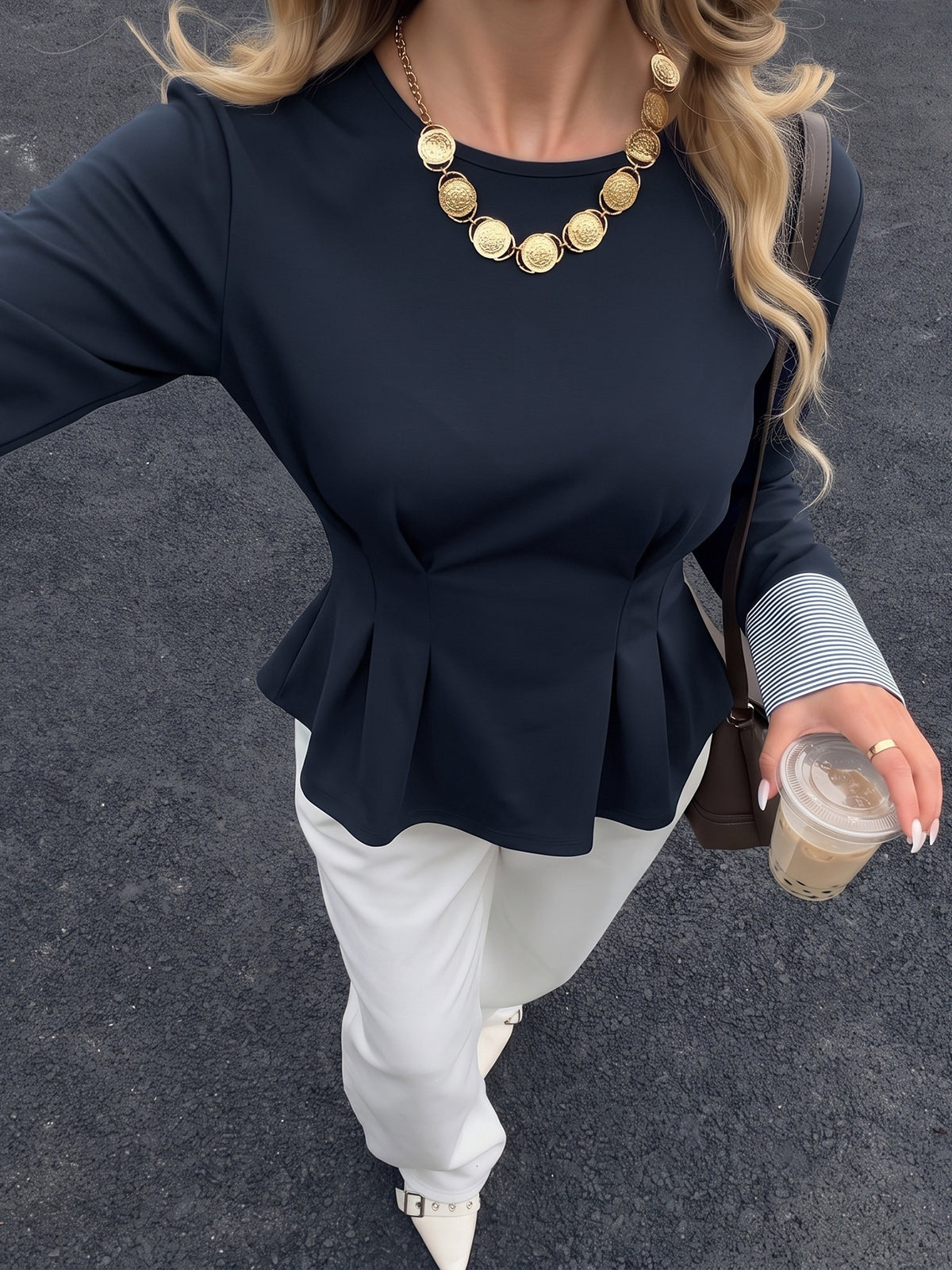 A woman wearing a navy blue long-sleeve peplum top, white pants, and a chunky gold disc necklace, holding an iced coffee on a dark asphalt surface.