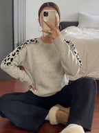 A woman sitting on a wooden floor in a bedroom setting, wearing a light beige crewneck sweater with black leopard print details on the shoulders, paired with black pants and white slippers.