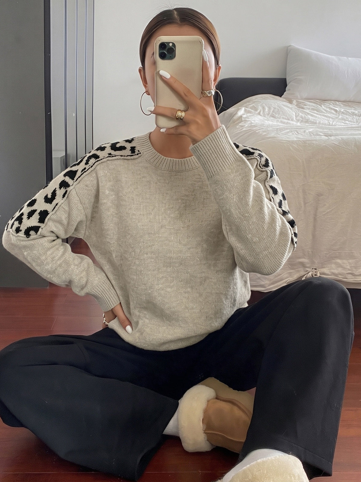A woman sitting on a wooden floor in a bedroom setting, wearing a light beige crewneck sweater with black leopard print details on the shoulders, paired with black pants and white slippers.