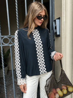 A woman wearing a navy blue long-sleeve blouse with white lace trim down the front and on the sleeves, paired with white pants and holding a brown mesh tote bag, standing in front of a building entrance.