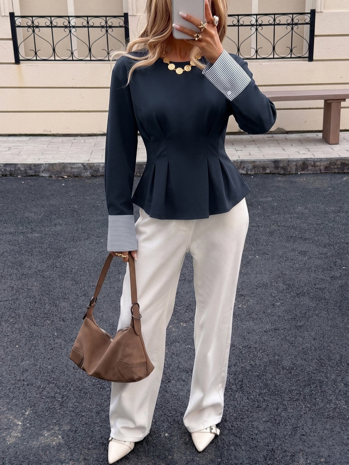A woman wearing a dark navy long-sleeve peplum top with light gray cuffs, white wide-leg trousers, and white pointed shoes, holding a brown leather shoulder bag. She is standing outdoors on a paved street in front of a building.