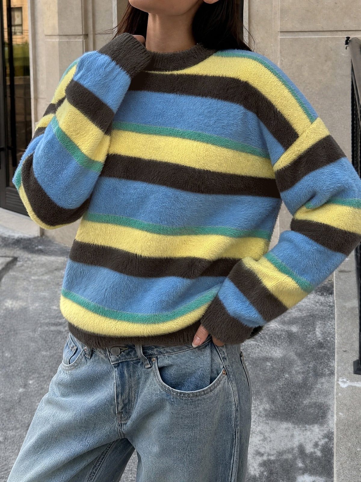 A person wearing a fuzzy knit sweater with horizontal stripes in light blue, yellow, dark brown, and thin teal lines, paired with light blue jeans.