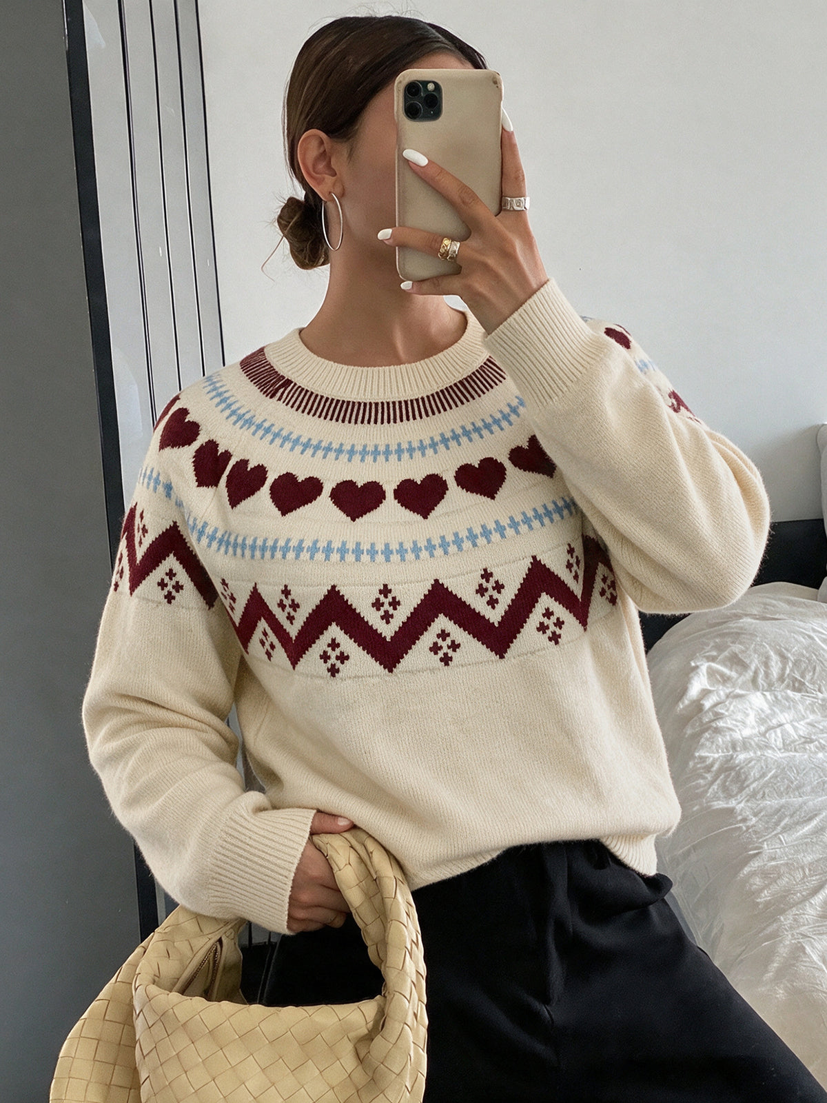 A woman takes a mirror selfie wearing a cream-colored knit sweater with a Fair Isle pattern featuring dark red hearts and geometric zigzags, paired with black pants and holding a woven beige bag.