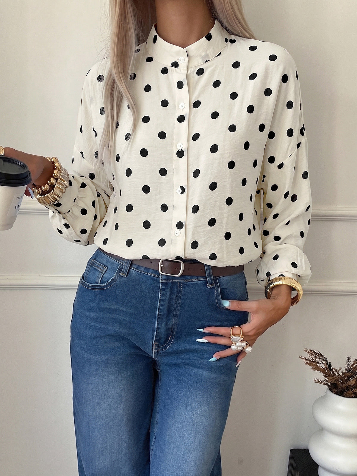 A woman wearing a cream-colored long-sleeve button-up blouse with black polka dots, paired with blue jeans and a brown belt, holding a coffee cup.