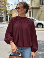 A woman wearing a long-sleeve burgundy blouse with a high gathered neckline and voluminous sleeves, paired with light blue jeans, standing outdoors on a city street.
