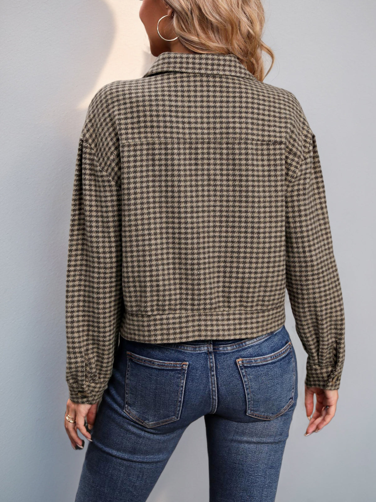 A woman shown from the back wearing a cropped, long-sleeve plaid jacket in brown and beige, paired with blue jeans, against a plain light gray wall.