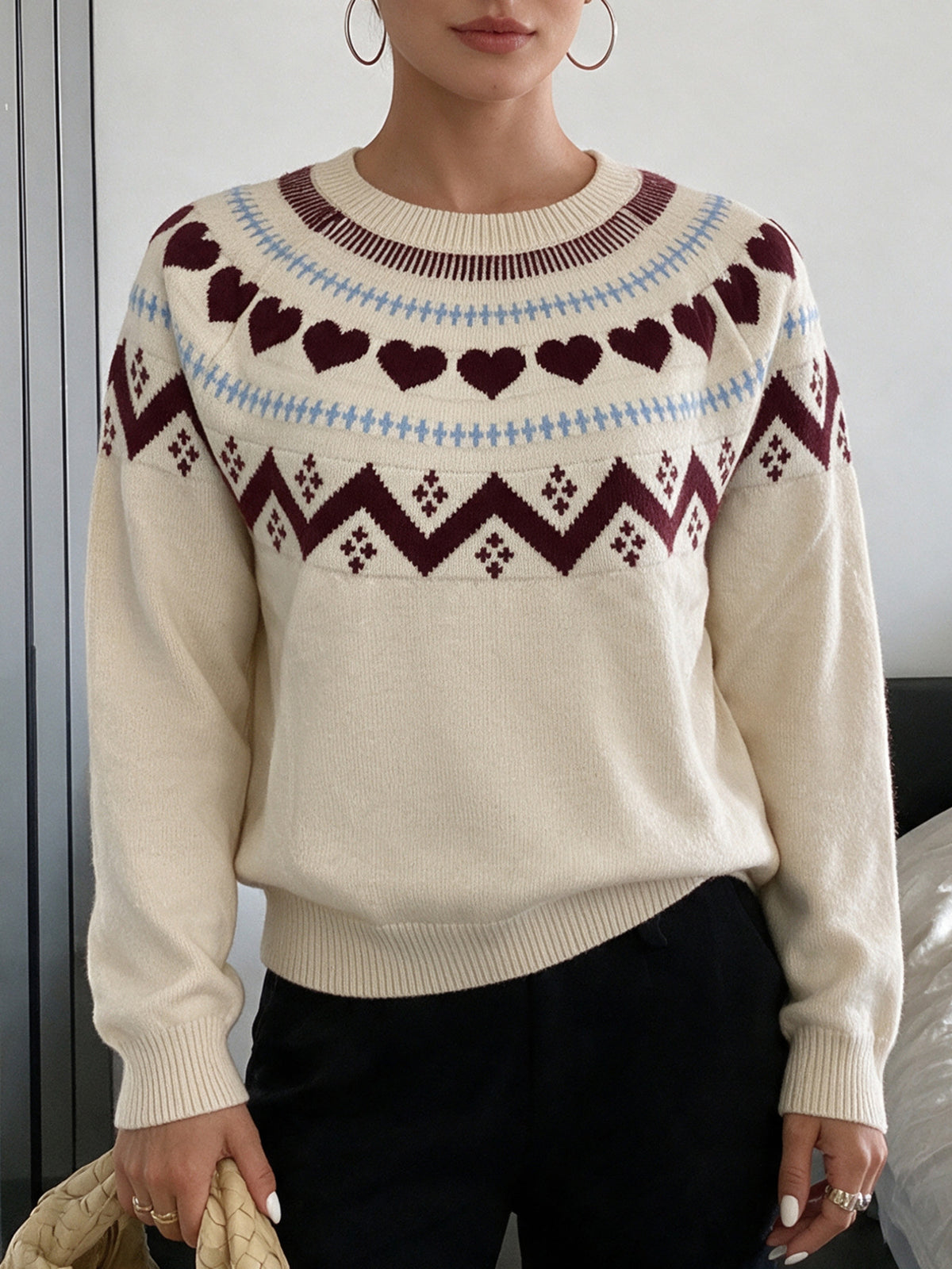 A woman wearing a cream-colored knit sweater with a Fair Isle pattern featuring dark red hearts, blue zigzags, and dark red geometric designs across the chest and shoulders, paired with black pants.