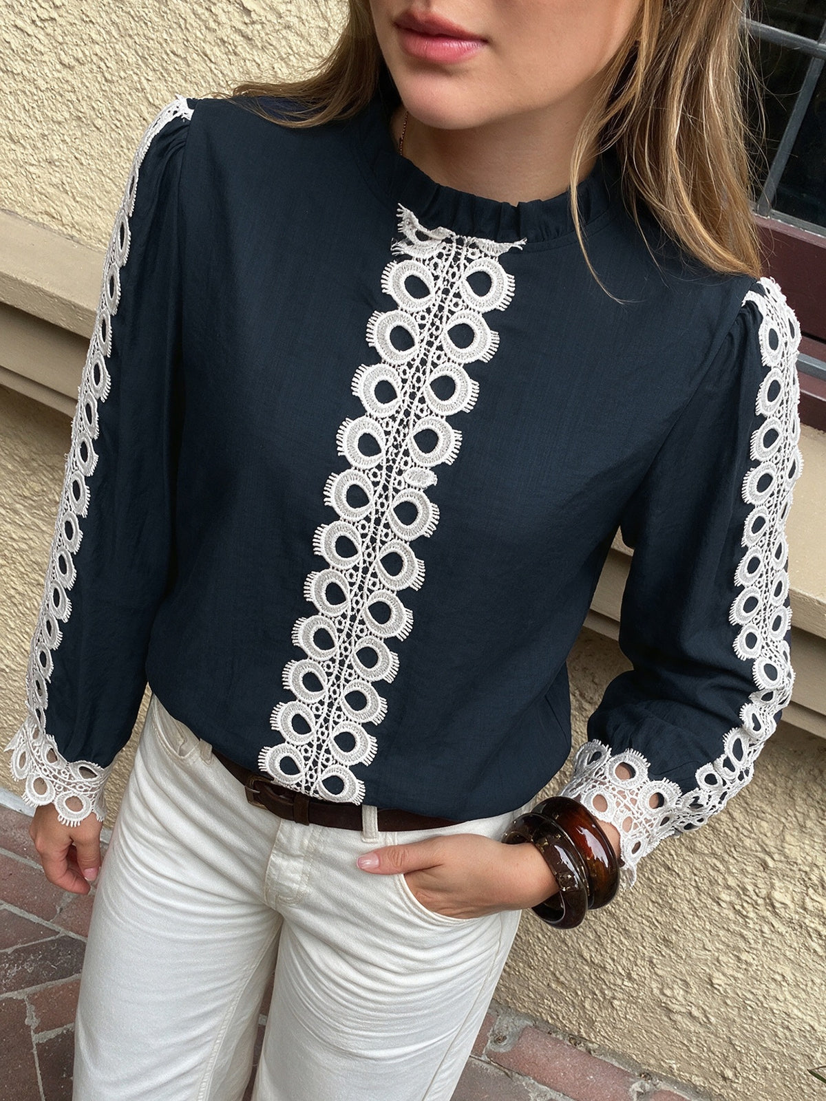 A woman wearing a navy blue long-sleeve blouse with white lace trim down the front and along the sleeves, paired with white pants.