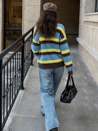 A person walking away on a city sidewalk wearing a fuzzy knit sweater with horizontal stripes in blue, yellow, and brown, paired with light blue jeans and a leopard-print beret, while carrying a black handbag.