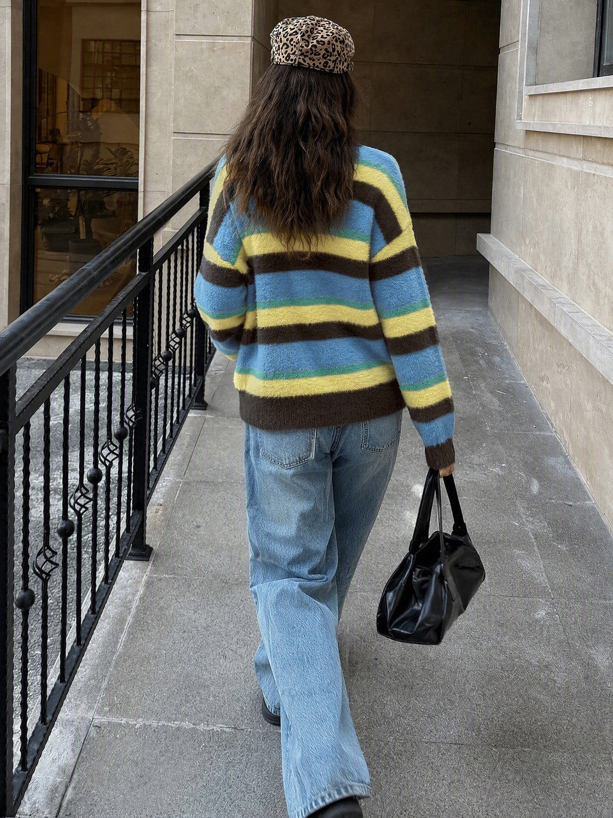 A person walking away on a city sidewalk wearing a fuzzy knit sweater with horizontal stripes in blue, yellow, and brown, paired with light blue jeans and a leopard-print beret, while carrying a black handbag.