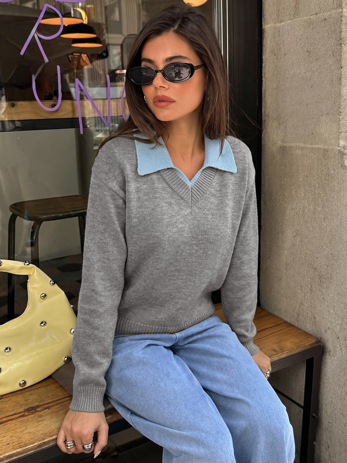 A woman wearing a gray knit sweater with a light blue collar, paired with light blue sweatpants and black sunglasses, sitting on a wooden bench.