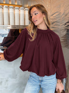 A woman wearing a burgundy long-sleeve blouse with a gathered high neck and voluminous sleeves, paired with light blue jeans, standing in a cafe-like environment.