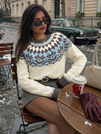 A woman sits at an outdoor cafe table wearing a cream knit sweater with a brown and light blue geometric pattern across the chest, paired with a gray mini skirt and dark sunglasses. A coffee cup, gloves, and a handbag are on the wooden table in front of her, with a street scene and a vintage green car in the background.