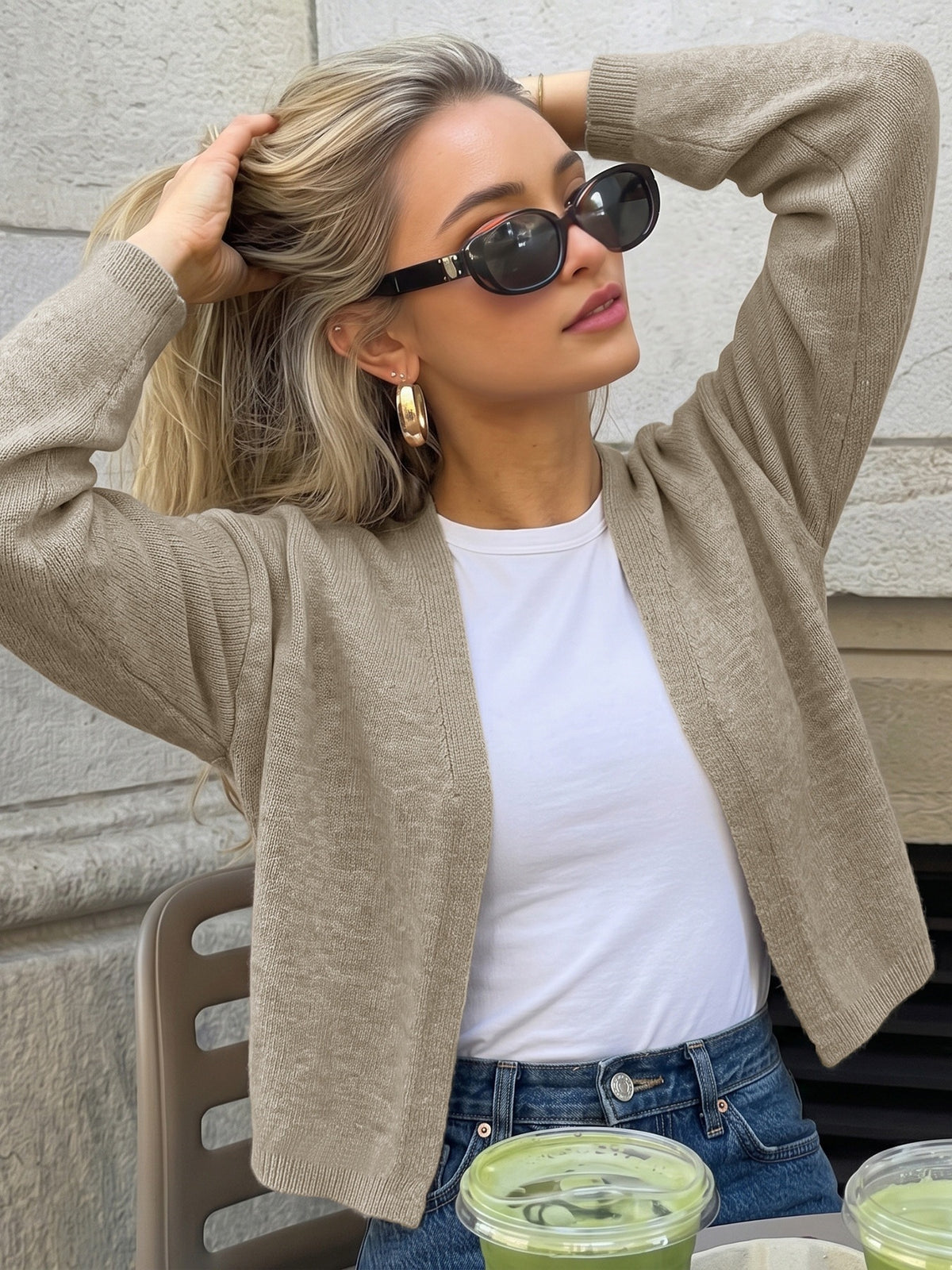A woman wearing a beige knit cardigan over a white t-shirt and blue jeans, styled with black sunglasses and gold hoop earrings, posing outdoors against a stone wall.