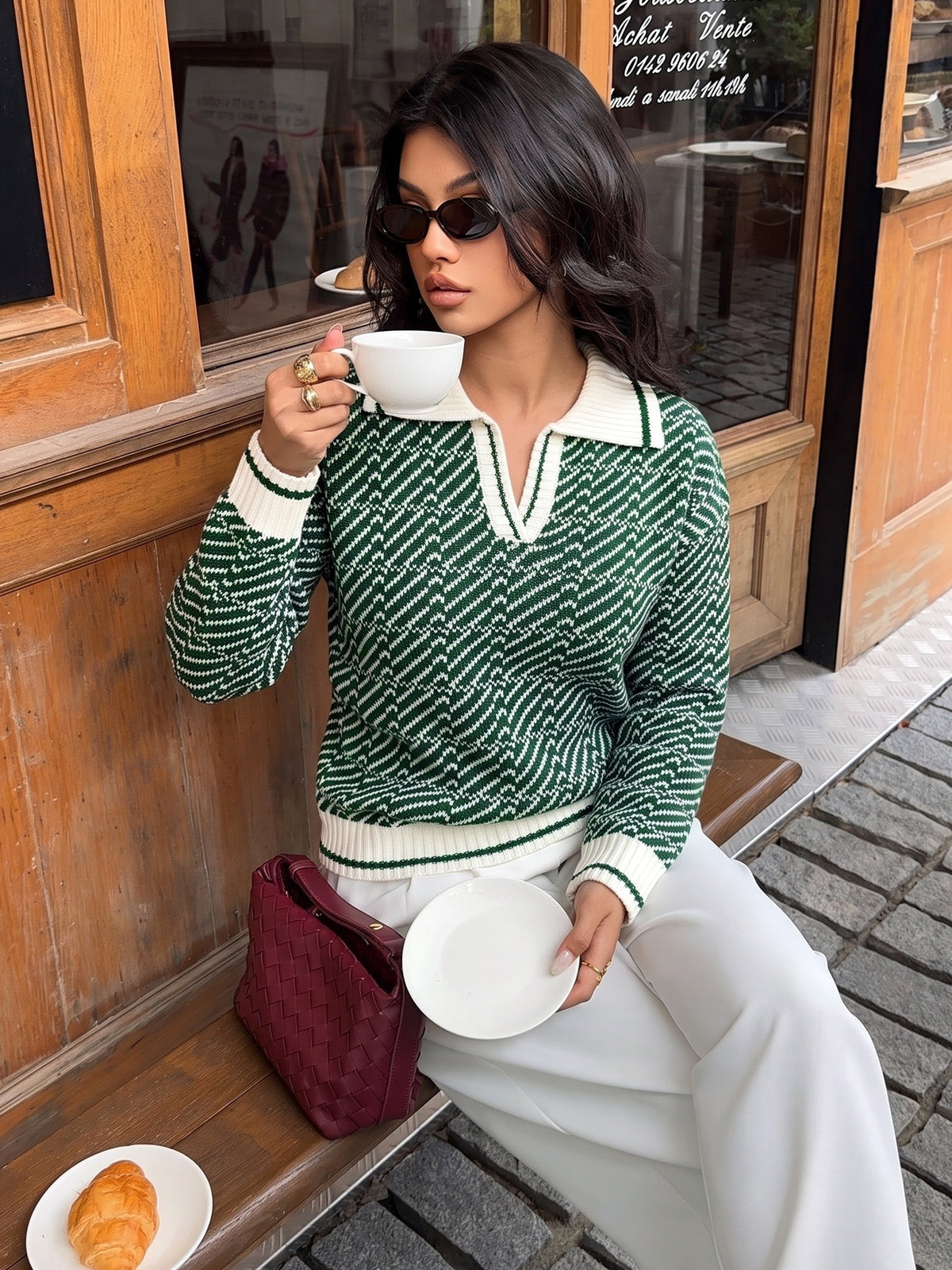 A woman wearing a green and white patterned knit polo sweater sits on a wooden bench outside a shop, holding a white coffee cup and saucer. She is wearing white trousers and a burgundy quilted handbag.
