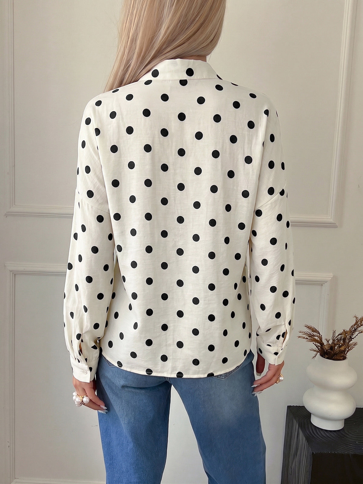 A woman shown from the back wearing a white long-sleeve blouse with black polka dots and blue jeans, standing indoors against a light paneled wall.