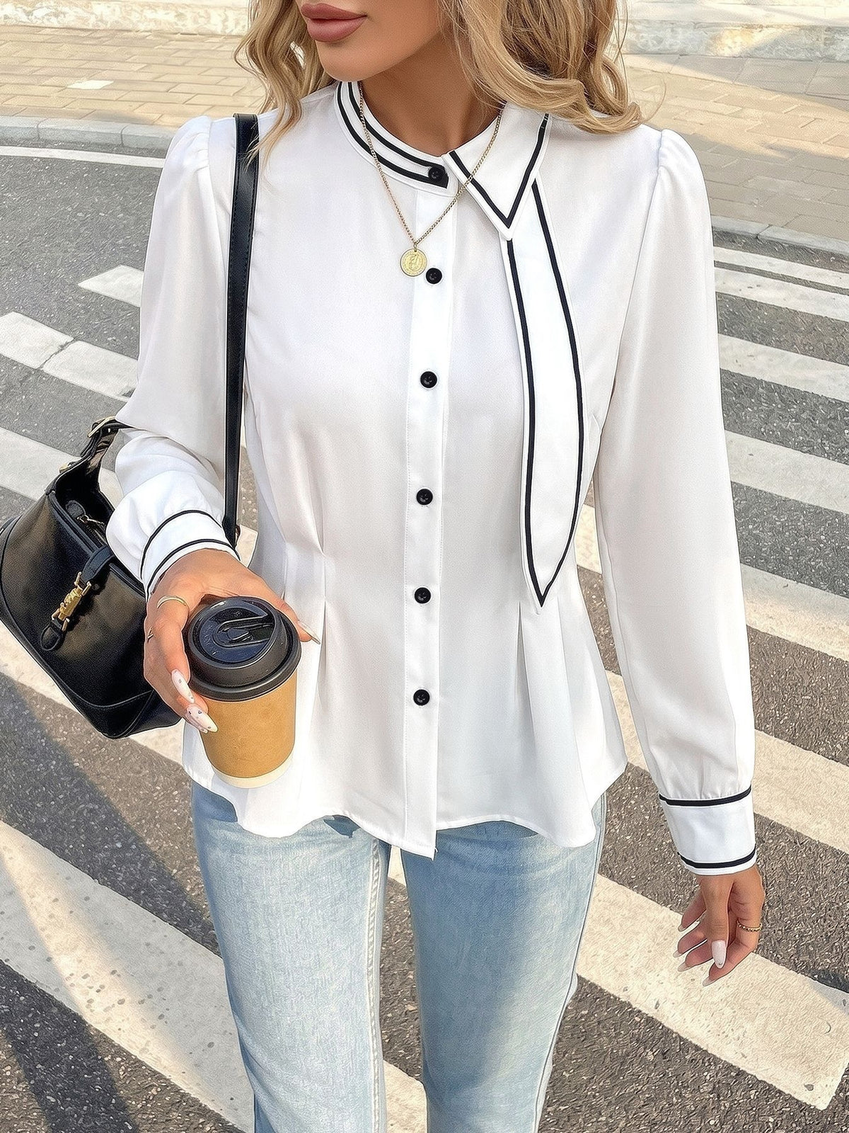 A woman wearing a white long-sleeve blouse with black piping and black buttons, paired with light blue jeans and carrying a black shoulder bag while holding a coffee cup on a street crosswalk.