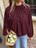 A woman wearing a long-sleeve, high-neck, gathered blouse in deep burgundy, paired with light blue jeans and holding a bouquet of white flowers.