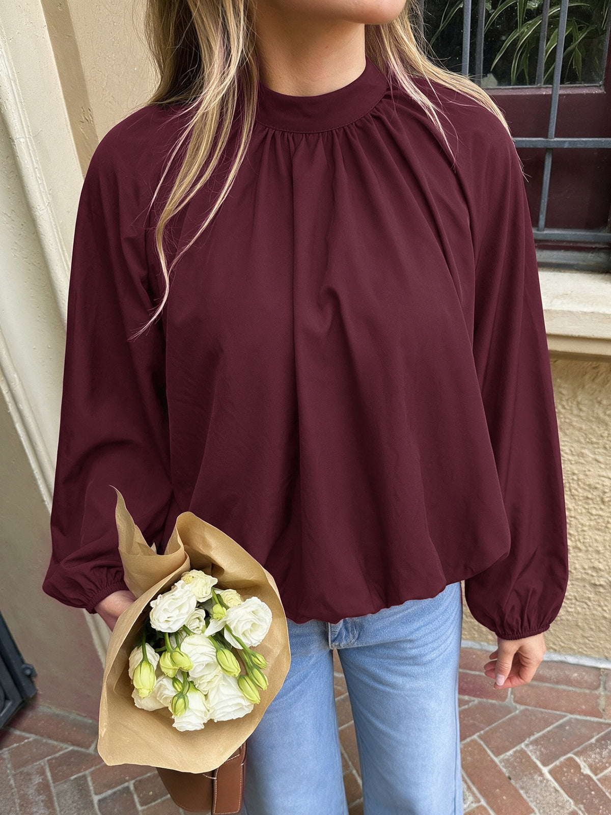 A woman wearing a long-sleeve, high-neck, gathered blouse in deep burgundy, paired with light blue jeans and holding a bouquet of white flowers.