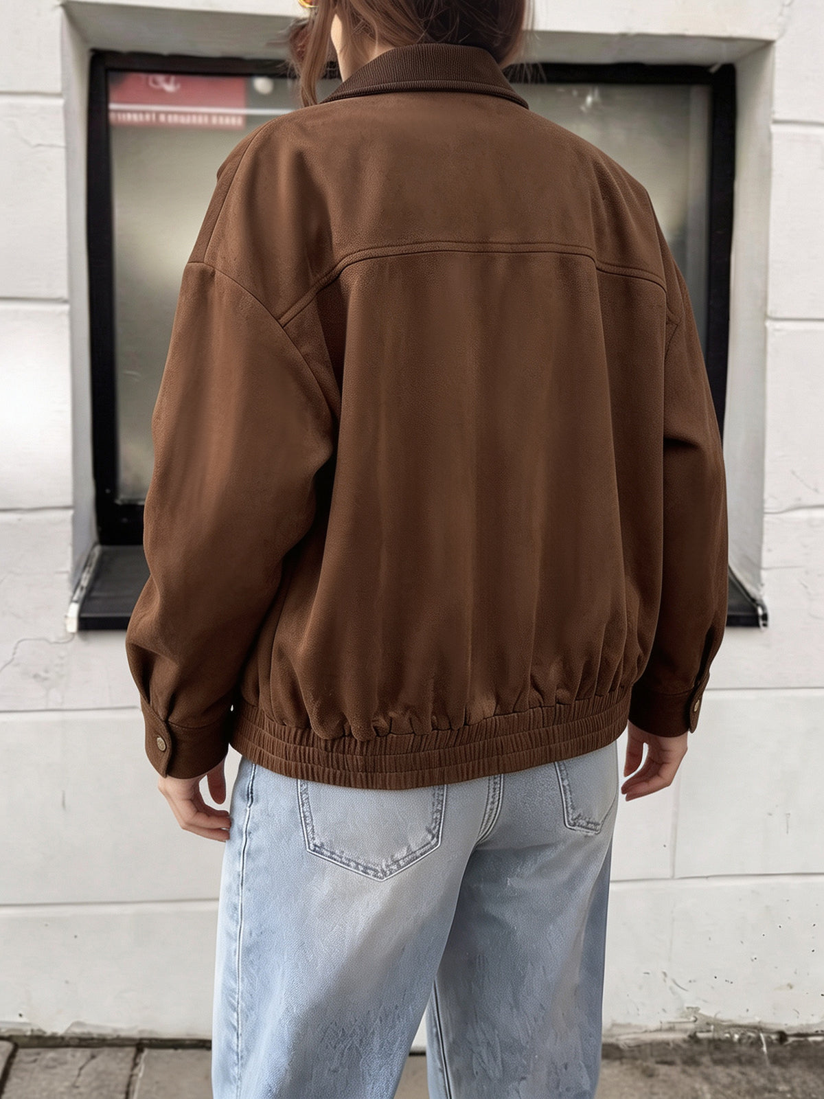 A person viewed from behind wearing a brown oversized jacket and light blue jeans, standing in front of a white brick wall.