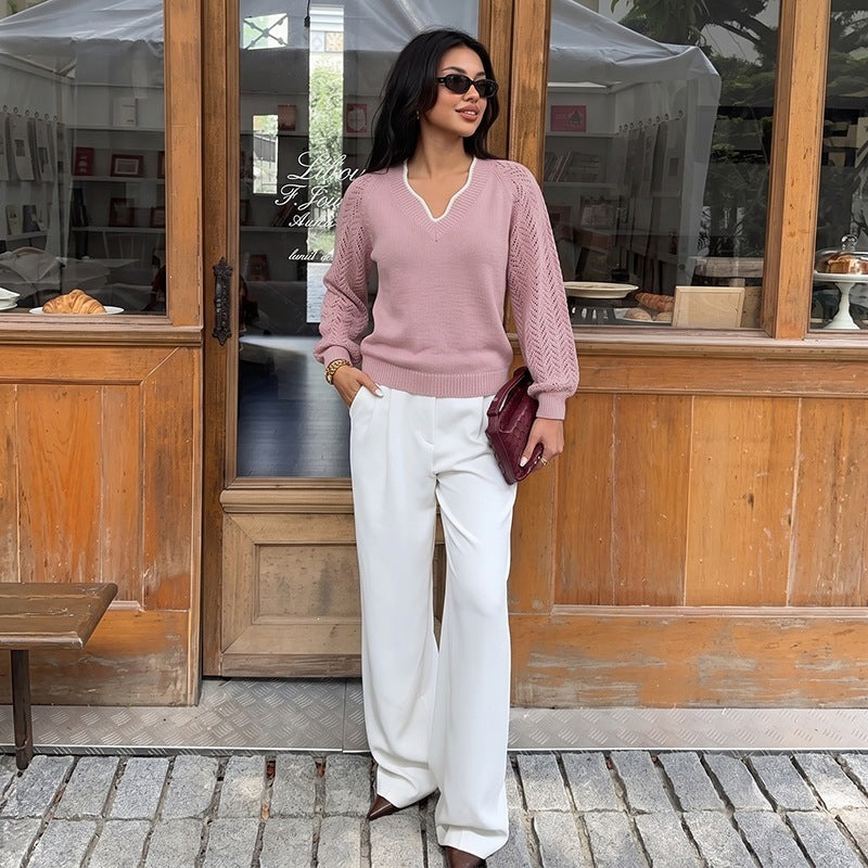 A woman stands in front of a rustic wooden storefront wearing a dusty pink knit sweater with a V-neck, white wide-leg pants, and holding a dark burgundy clutch.