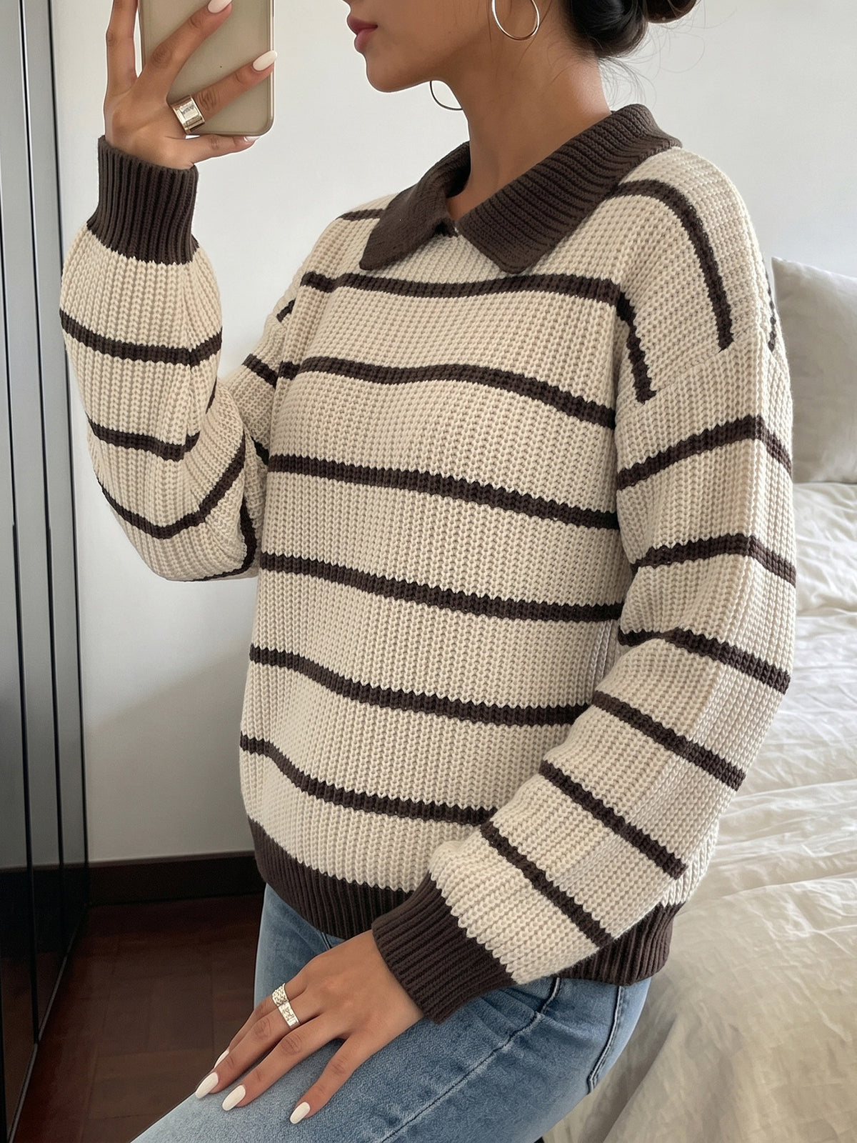 A woman wearing a chunky knit sweater with horizontal dark brown stripes on an off-white base, paired with blue jeans, taking a mirror selfie in a bedroom setting.