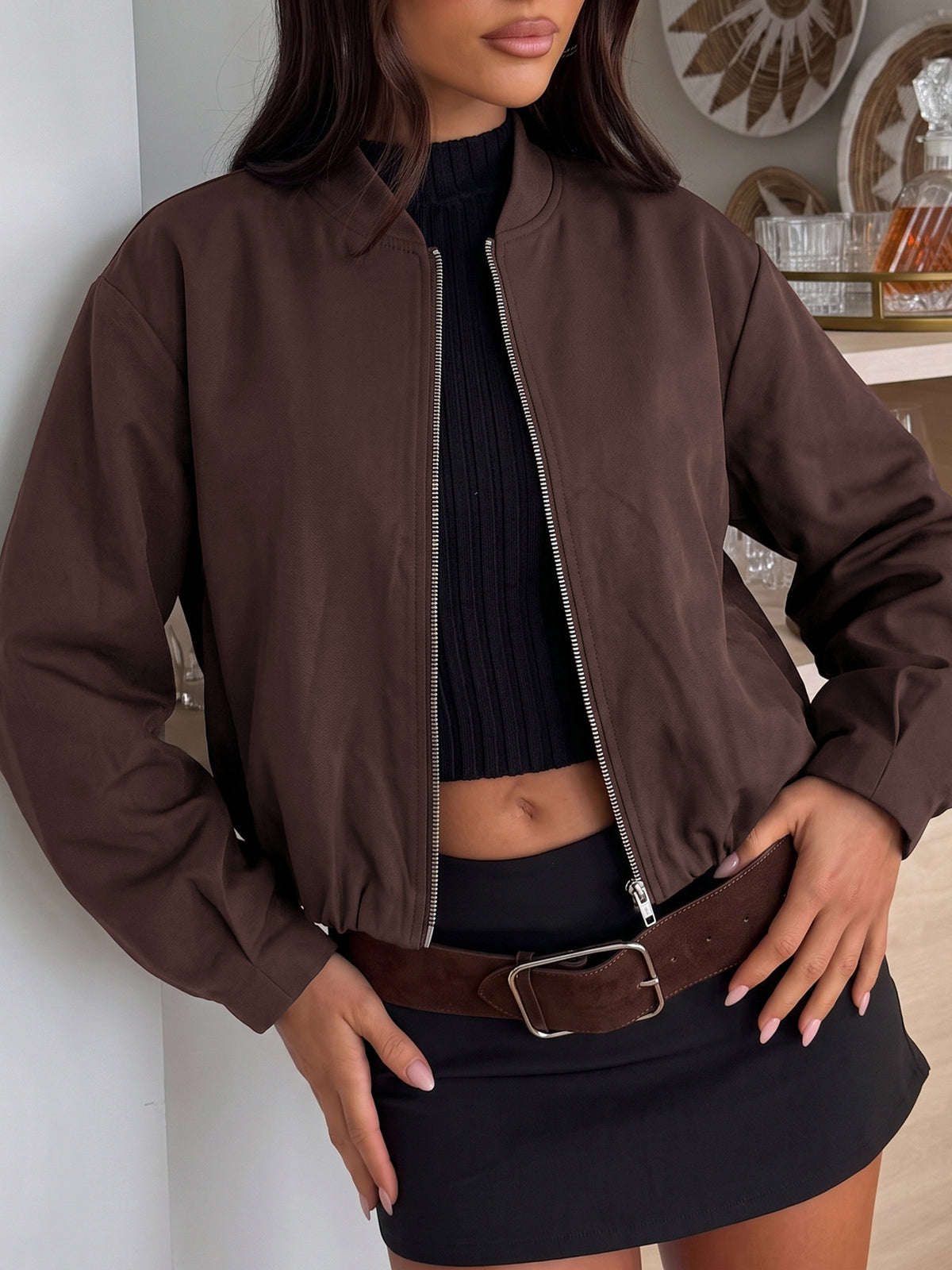 A woman wearing a brown zip-up bomber jacket over a black ribbed crop top and black mini skirt, with a brown belt, standing indoors against a light wall.