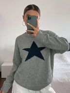 A woman takes a mirror selfie wearing a heather gray knit sweater with a large dark navy blue star on the front, paired with white pants.