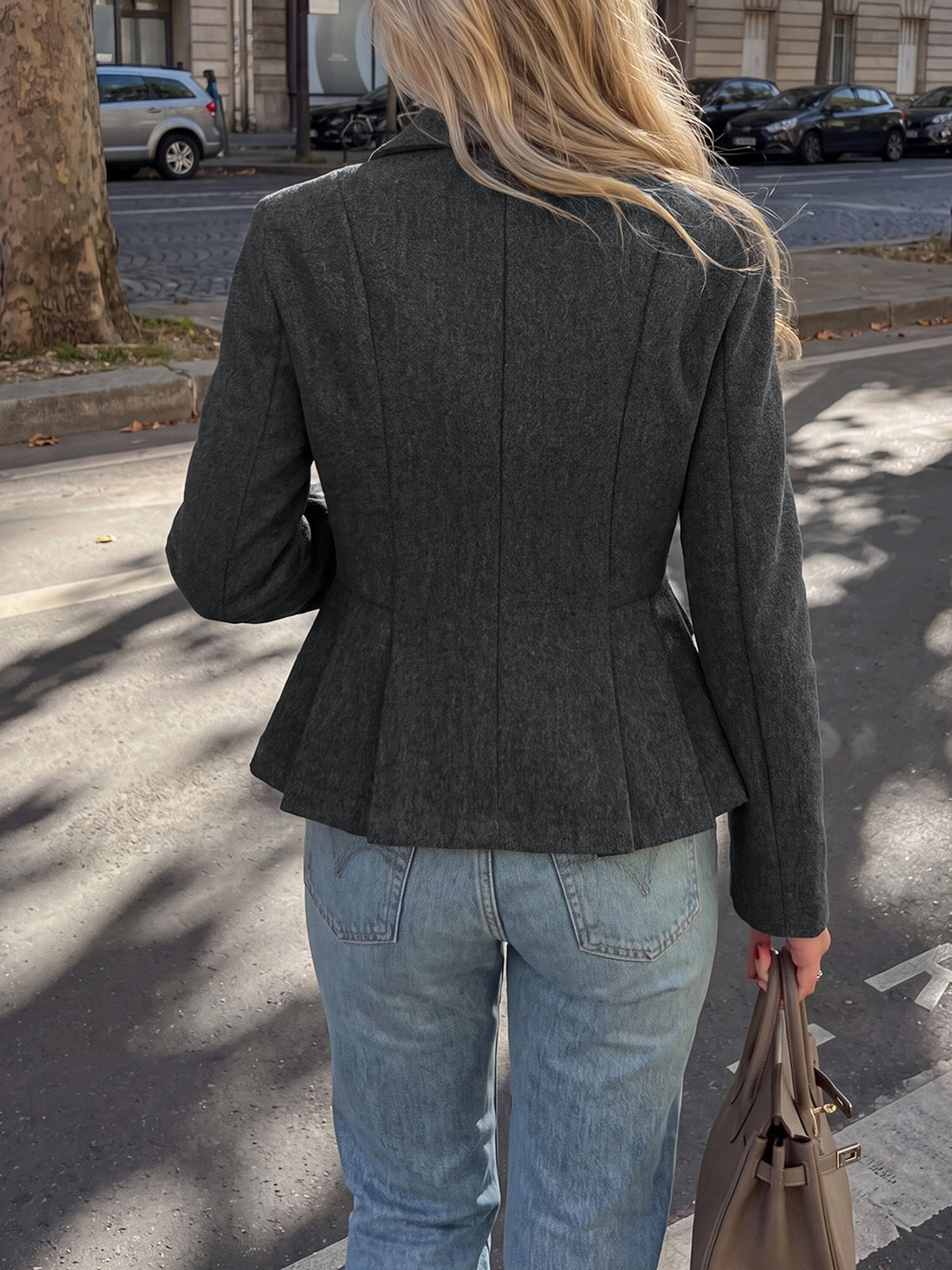 A woman viewed from behind wearing a dark gray textured peplum blazer and light blue jeans, holding a taupe handbag on an outdoor street.