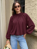 A woman wearing a burgundy long-sleeve blouse with a gathered high neck and voluminous sleeves, paired with light blue jeans, standing outdoors against a textured beige wall.