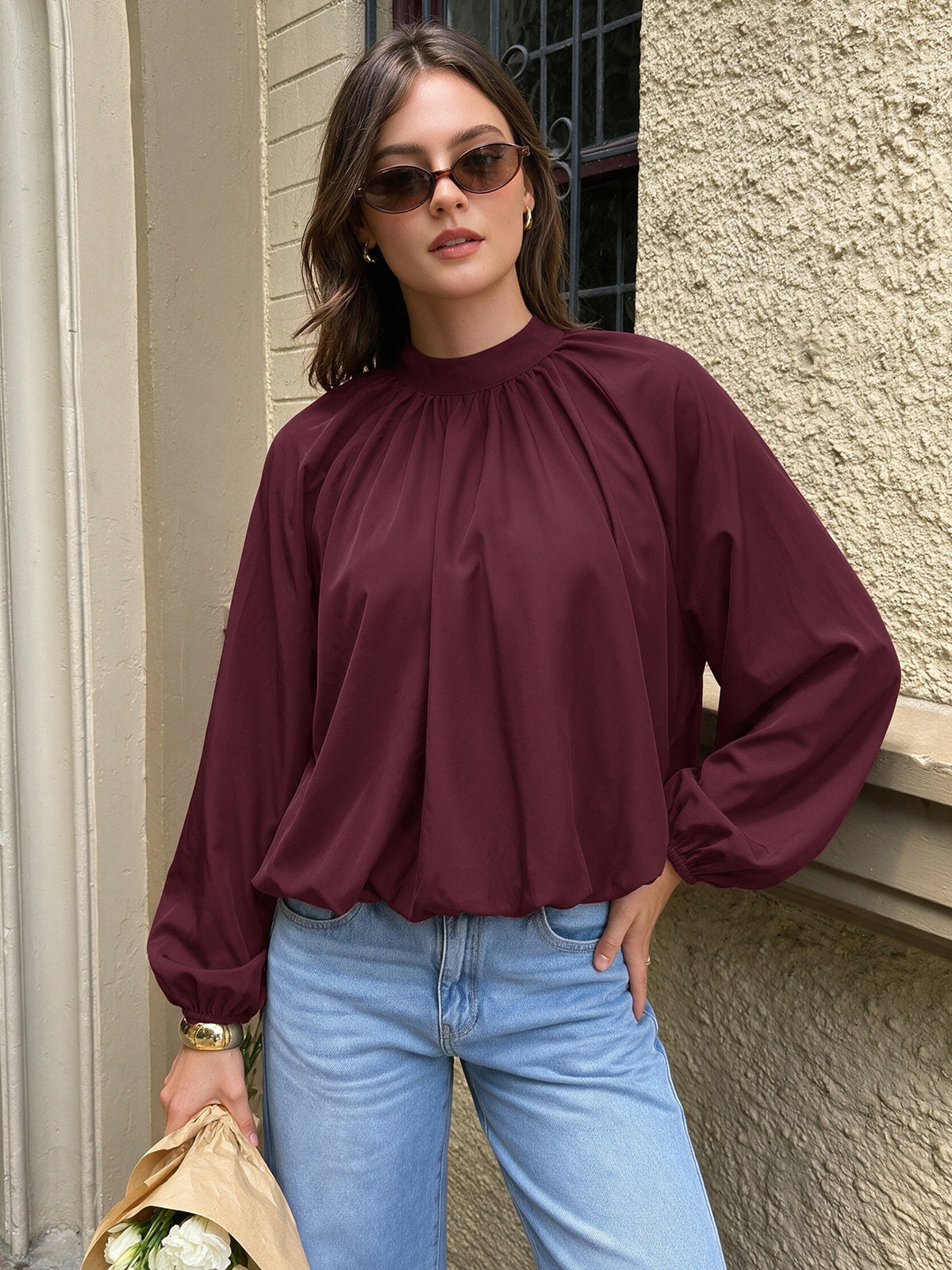A woman wearing a burgundy long-sleeve blouse with a gathered high neck and voluminous sleeves, paired with light blue jeans, standing outdoors against a textured beige wall.