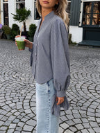 A woman wearing a blue and white gingham long-sleeve blouse with a shawl collar and gathered sleeves, paired with light-wash denim jeans, standing outdoors on a cobblestone street and holding a green drink.