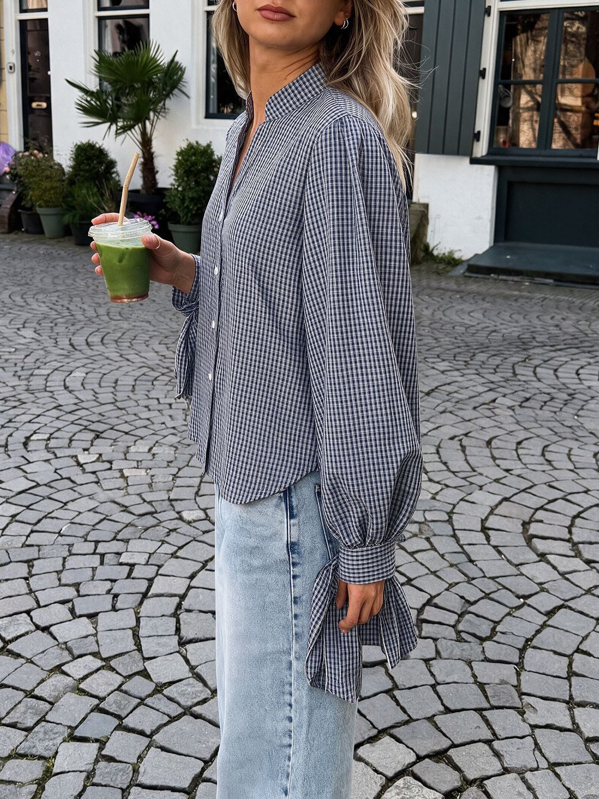 A woman wearing a blue and white gingham long-sleeve blouse with a shawl collar and gathered sleeves, paired with light-wash denim jeans, standing outdoors on a cobblestone street and holding a green drink.
